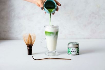 Matcha Green Tea Latte In Between a Chasen Matcha Whisk, a Chashuku Matcha Spoon, And A Tin Of Kineta Matcha Tea